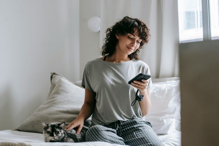 A female pet owner sitting on a bed looking at types of pet insurance on her phone while petting her cat.
